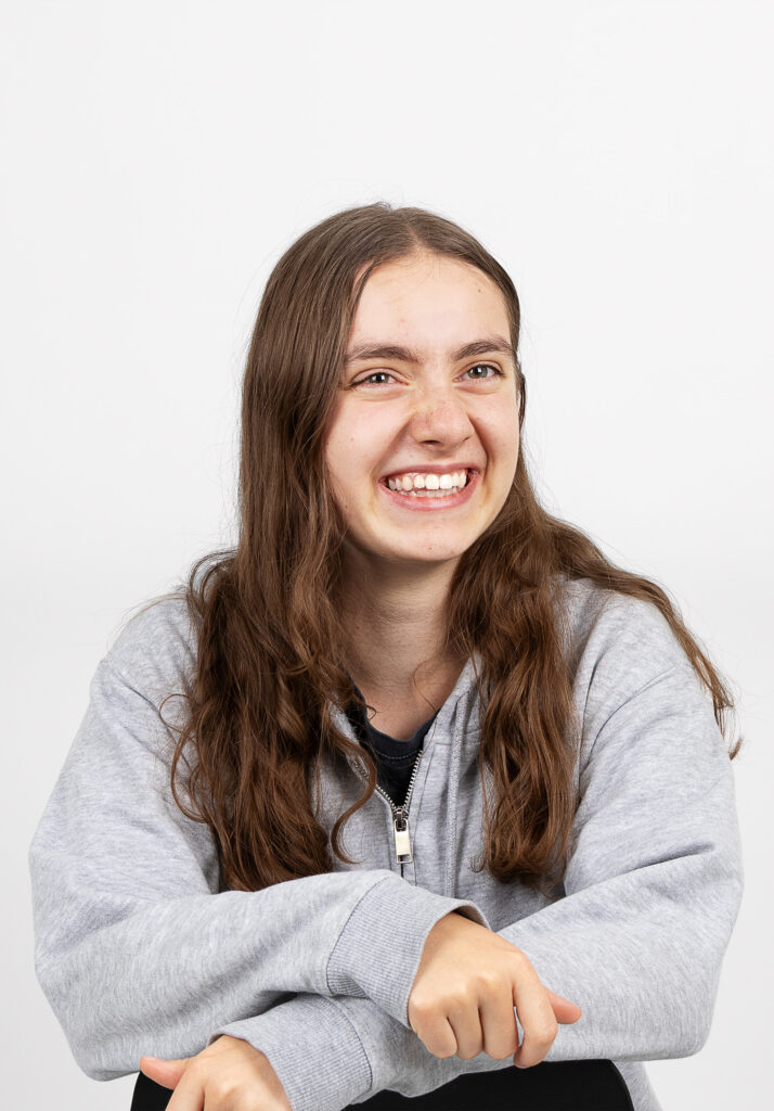 Eine junge Person mit langen braunen Haaren, die einen grauen Kapuzenpullover mit Reißverschluss trägt, lächelt breit und blickt leicht zur Seite vor einem schlichten weißen Hintergrund.