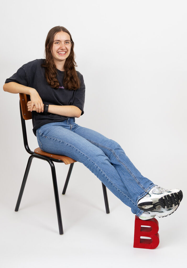 Eine Person mit langen braunen Haaren, die ein dunkles Hemd, blaue Jeans und Turnschuhe trägt, sitzt auf einem Stuhl, die Beine ausgestreckt und die Füße auf einem großen roten Buchstaben "B" ruhend. Der Hintergrund ist einfarbig weiß.
