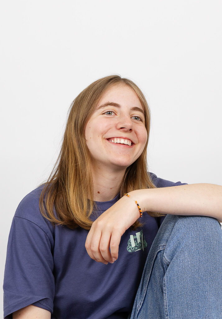 Eine junge Person mit langem, hellbraunem Haar, die ein blaues T-Shirt und Jeans trägt, sitzt mit einem angehobenen Knie und lächelt, während sie vor einem schlichten weißen Hintergrund leicht zur Seite schaut.