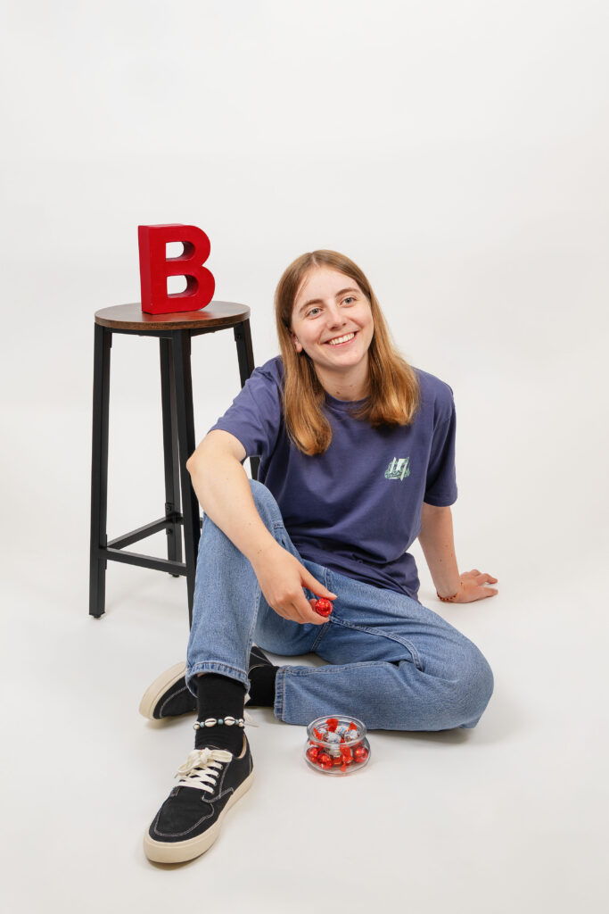 Eine Person mit langen Haaren, blauem T-Shirt und Jeans, sitzt lächelnd auf dem Boden und hält eine rote Kapsel in der Hand. Daneben steht ein Hocker mit einem großen roten Buchstaben "B" darauf und einer Schale mit weiteren roten Kapseln.