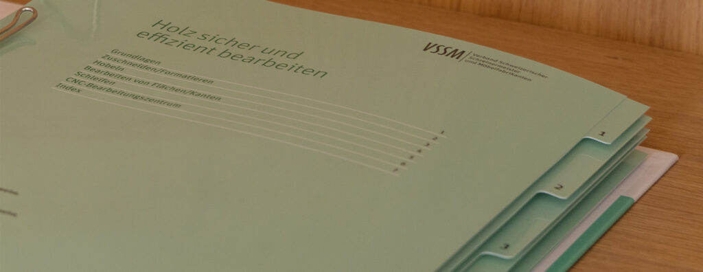 Ein grüner Ordner mit der Aufschrift „Holz sicher und effizient bearbeiten“ mit dem VSSM-Logo auf einer Holzoberfläche. Auf dem Cover sind verschiedene Abschnitte wie Grundlagen, Holzarten und Arbeitstechniken aufgeführt.