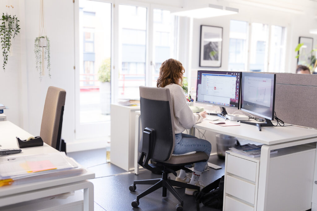Eine Person mit lockigem Haar sitzt an einem Schreibtisch in einem modernen Büro und arbeitet an zwei Monitoren.