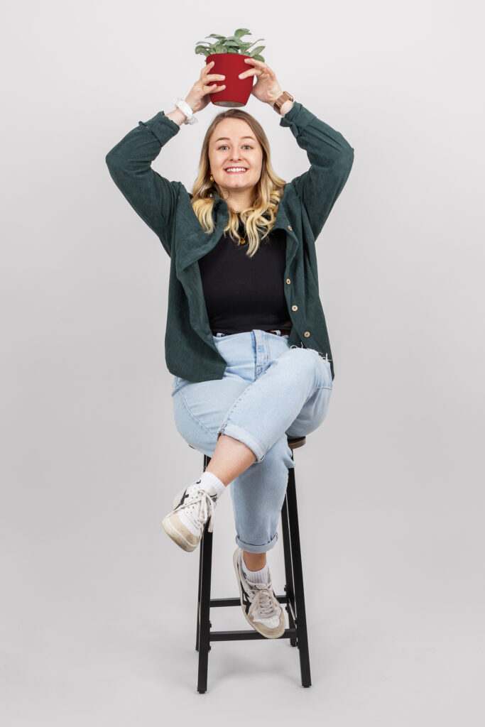 Eine Person mit langen Haaren, die eine grüne Jacke, ein schwarzes Hemd, hellblaue Jeans und weiße Turnschuhe trägt, sitzt auf einem Hocker, hält eine kleine Topfpflanze über dem Kopf und lächelt vor einem schlichten Hintergrund.