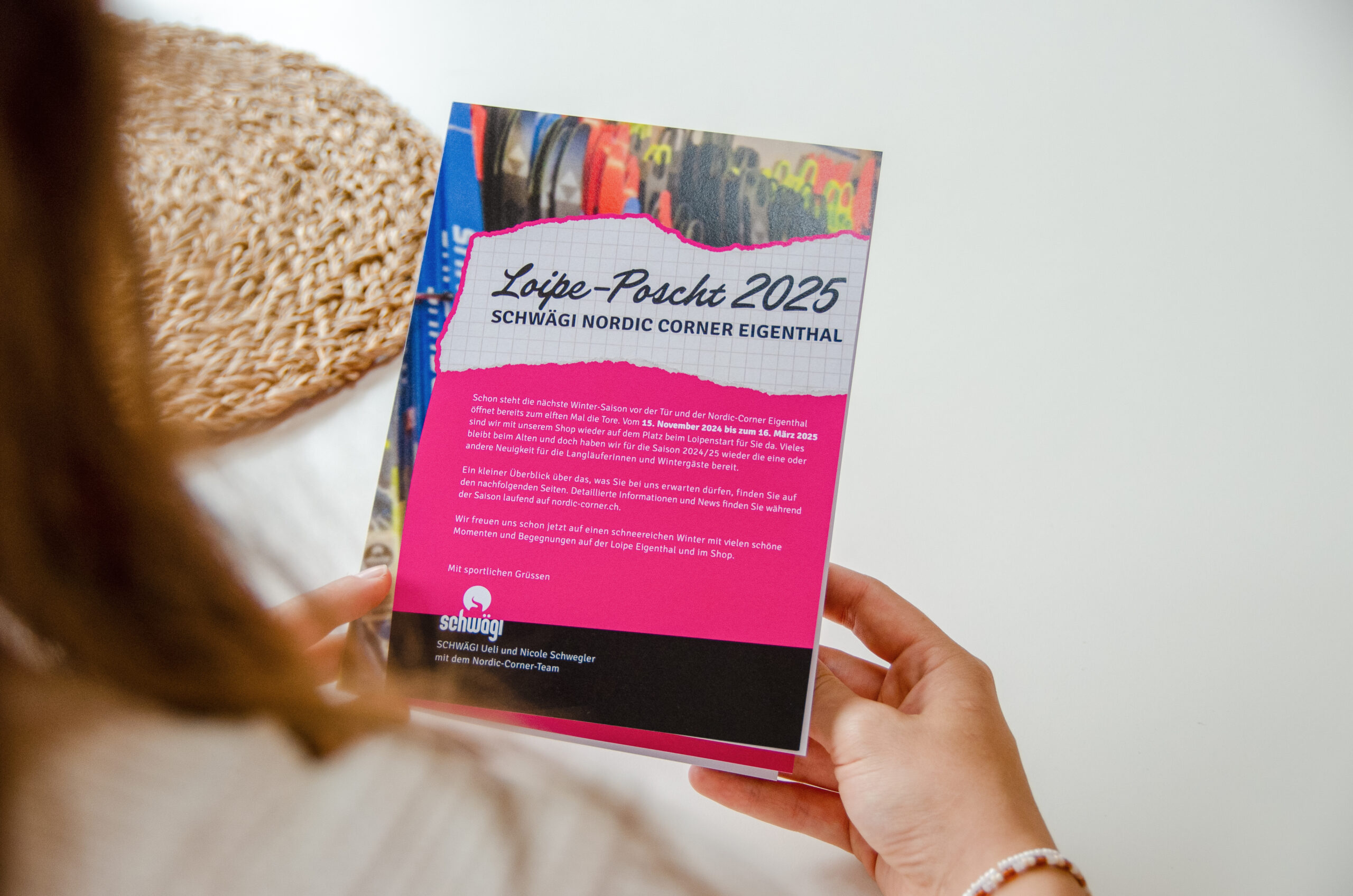 Eine Person hält eine Broschüre mit dem Titel „Löipe-Poscht 2025“ mit einem farbenfrohen Cover und einem Text, der den Schwägi Nordic Corner Eigenthal beschreibt. Die Broschüre enthält Sport- und Veranstaltungsinformationen. Im Hintergrund ist ein geflochtener Korb teilweise sichtbar.