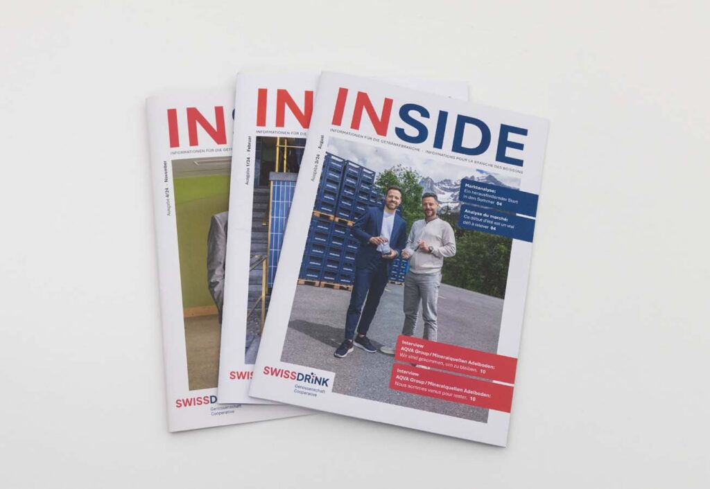 Drei Ausgaben des Magazins „INSIDE“ liegen gestapelt. Das Cover zeigt zwei Männer mit Getränken vor gestapelten Kisten im Freien. Die Überschrift ist in Rot und Blau gehalten.