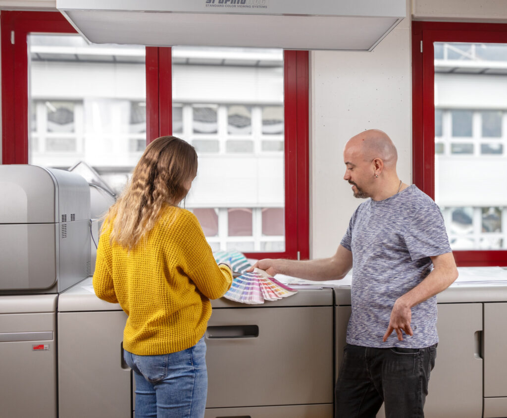 Ein Mann und eine Frau stehen vor einem großen Drucker. Der Mann im grauen Hemd gestikuliert mit Tatendrang auf das Farbmusterbuch, das die Frau im gelben Pullover in der Hand hält. Sie befinden sich in einem hell erleuchteten Raum mit rot gerahmten Fenstern.
