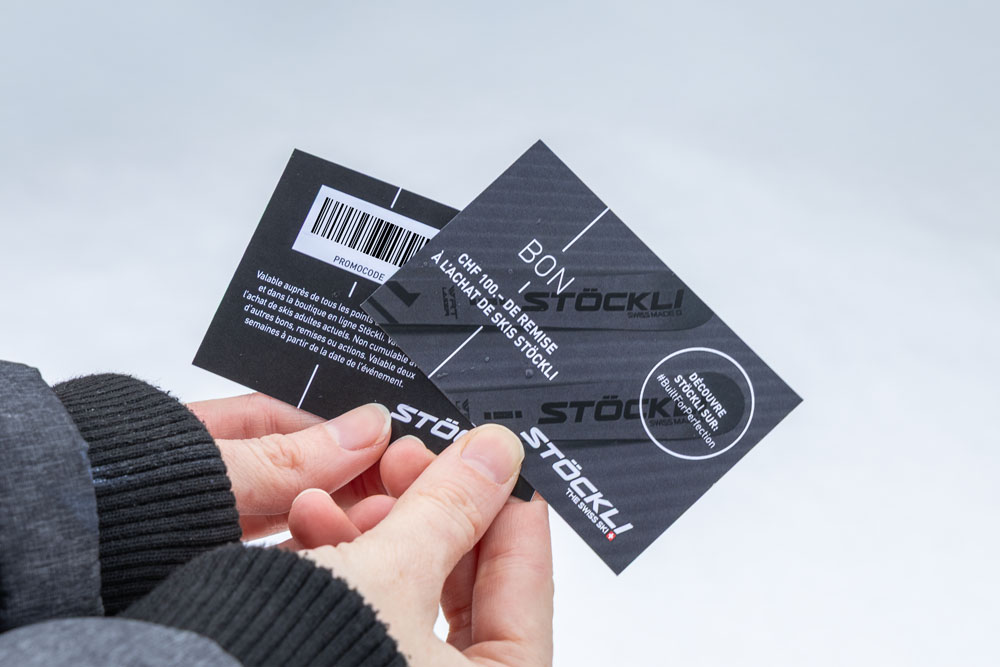 Eine Person hält zwei schwarze Geschenkkarten mit weißem Text und dem Markennamen „Stöckli“. Die Karten sind mit einem Barcode und Werbeinformationen versehen. Die Person trägt eine dunkle Jacke; der Hintergrund ist hellgrau.
