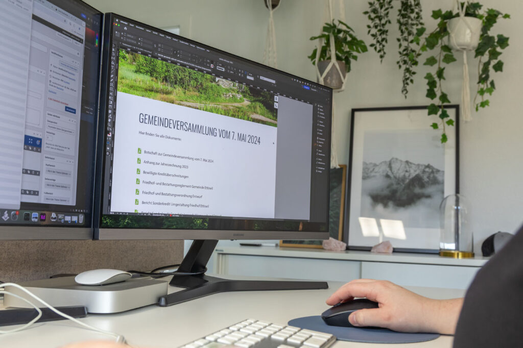 Eine Person arbeitet an einem Desktop-Computer mit einem großen Monitor, auf dem ein Dokument mit dem Titel „GEMEINDEVERSAMMLUNG VOM 7. MAI 2024“ angezeigt wird. Der Arbeitsplatz ist mit Pflanzen und einem gerahmten Bergfoto dekoriert.