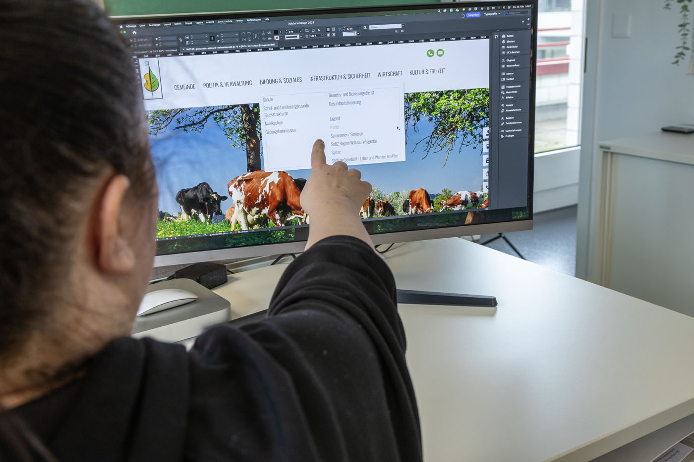 Eine Person mit dunklem Haar, die den Blick abwendet, zeigt auf einen Desktop-Monitor, auf dem eine Webdesign-Oberfläche angezeigt wird. Der Bildschirm zeigt ein grünes Menü und eine ländliche Szene mit Kühen. Der Arbeitsbereich ist hell dank des Tageslichts eines nahegelegenen Fensters.