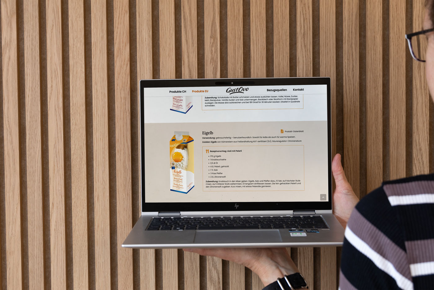 Eine Person hält einen Laptop in der Hand, auf dem eine Webseite über das Produkt „Eigelb“ mit einem Kartonbild auf einem Holzlattenhintergrund angezeigt wird. Das Gesicht der Person ist nicht sichtbar.