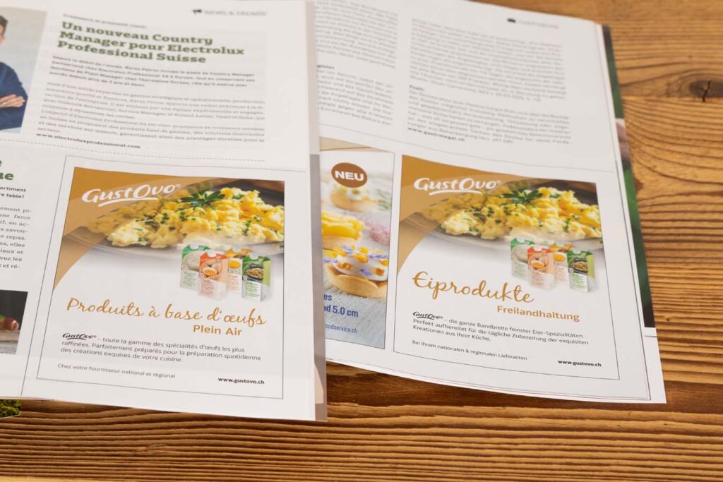 Aufgeschlagenes Magazin auf einer Holzoberfläche mit zwei Anzeigen für GustOvo-Eierprodukte. Die Anzeigen zeigen Bilder von Eierspeisen mit sichtbarem Markennamen und Produktverpackungen. Text in Französisch und Deutsch beschreibt die Produkte.