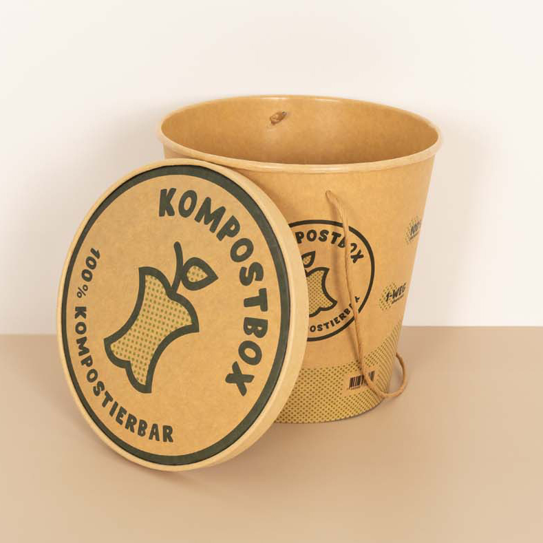 Eine braune Komposttonne mit der Aufschrift „KOMPOSTBOX“ und einer Apfelkerngrafik sowie dem Text „100% KOMPOSTIERBAR“ auf dem Deckel lehnt an der offenen Tonne.