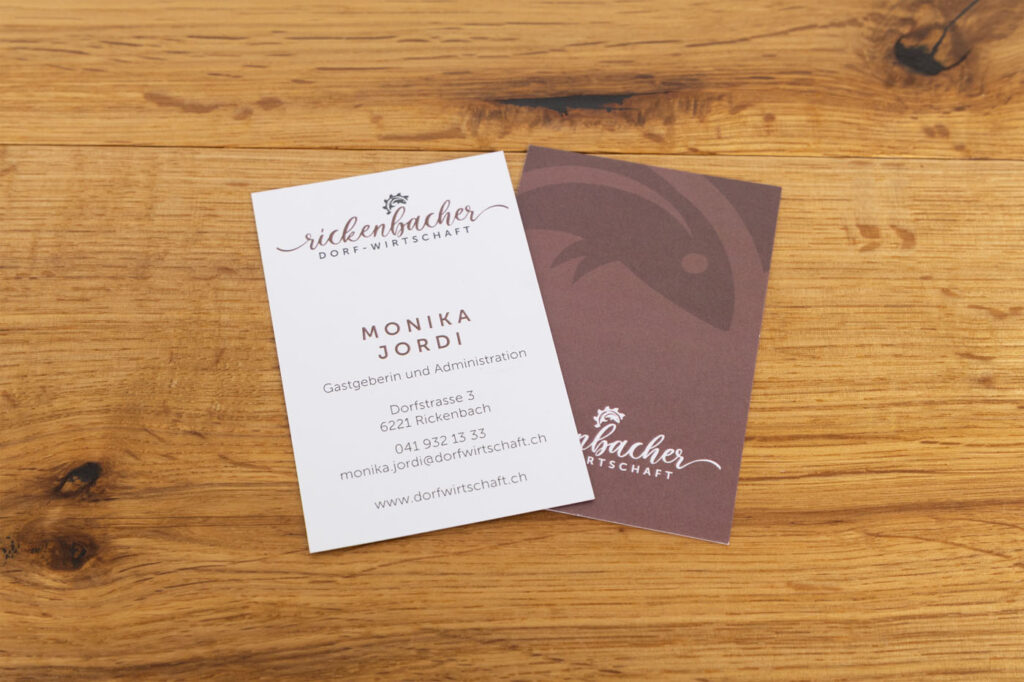 Zwei Visitenkarten auf Holzuntergrund. Die erste Karte zeigt die Kontaktdaten von Monika Jordi mit Adresse, Telefonnummer und E-Mail-Adresse. Die zweite Karte ist braun gestaltet und trägt das Logo der „Rickenbacher Dorfwirtschaft“.