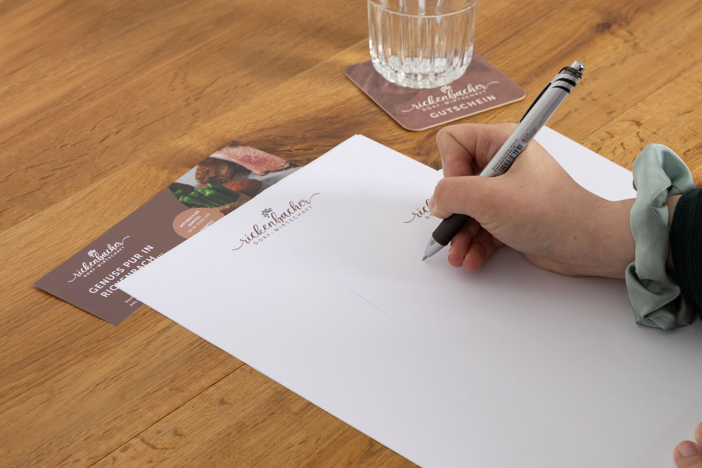 Eine Hand hält einen Stift und ist bereit, auf einem leeren Blatt Papier auf einem Holztisch zu schreiben. Daneben liegen Werbeflyer mit Text. Ein Glas Wasser steht auf einem Untersetzer daneben.
