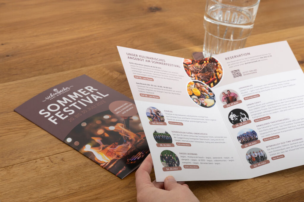 Eine Person hält eine Broschüre auf einem Holztisch. Auf dem Cover steht „Sommer Festival 20.–28.5.23“ mit einem Feuermotiv. Im Inneren befinden sich Fotos, Textdetails und ein QR-Code. Daneben steht ein Glas Wasser.