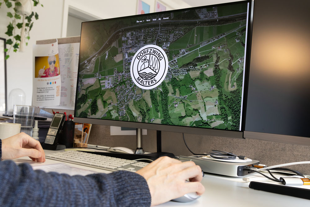 Eine Person bedient eine Computermaus. Auf einem großen Monitor ist eine Landschaftsansicht aus der Luft und in der Mitte ein Logo mit dem Schriftzug „Dorfzwilling Balters“ zu sehen. Der Schreibtisch ist mit Papieren, einem Telefon und Büromaterial übersät.
