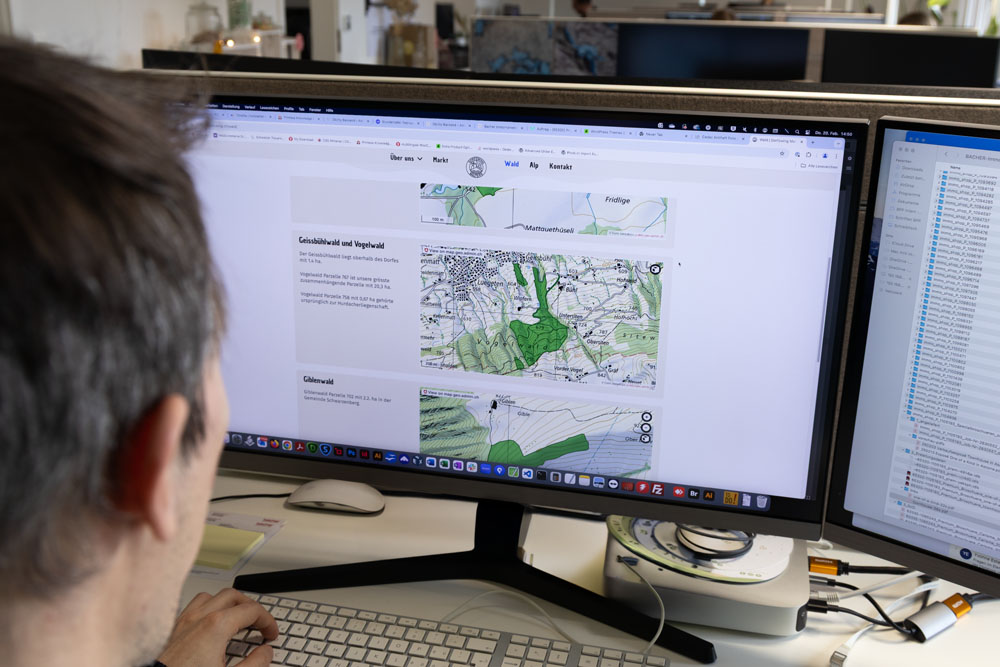 Eine Person sitzt an einem Schreibtisch und blickt auf einen großen Computerbildschirm mit Karten und Text. Der Bildschirm zeigt detaillierte geografische Karten und Informationen. Der Arbeitsbereich umfasst zwei Monitore und verschiedene Schreibtischgegenstände.