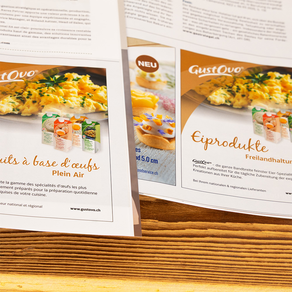 Aufgeschlagenes Magazin auf einer Holzoberfläche mit zwei Anzeigen für GustOvo-Eierprodukte. Die Anzeigen zeigen Bilder von Eierspeisen mit sichtbarem Markennamen und Produktverpackungen. Text in Französisch und Deutsch beschreibt die Produkte.