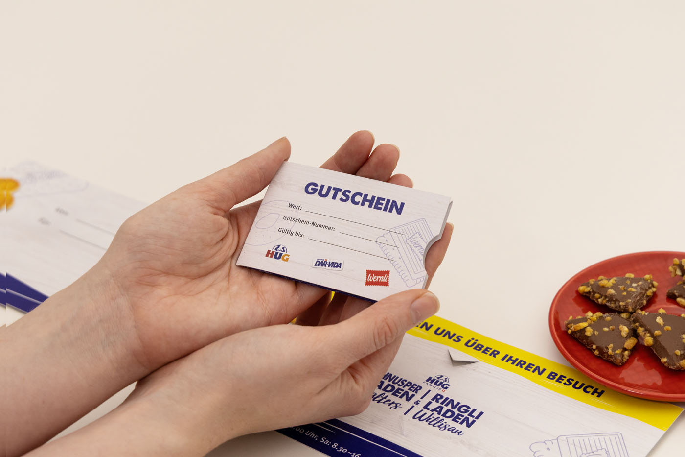 Zwei Hände halten einen Geschenkgutschein mit dem Aufdruck „Gutschein“. Im Hintergrund ist ein Tisch mit einigen Schokoladenstücken und Teilen anderer Papiergegenstände zu sehen.