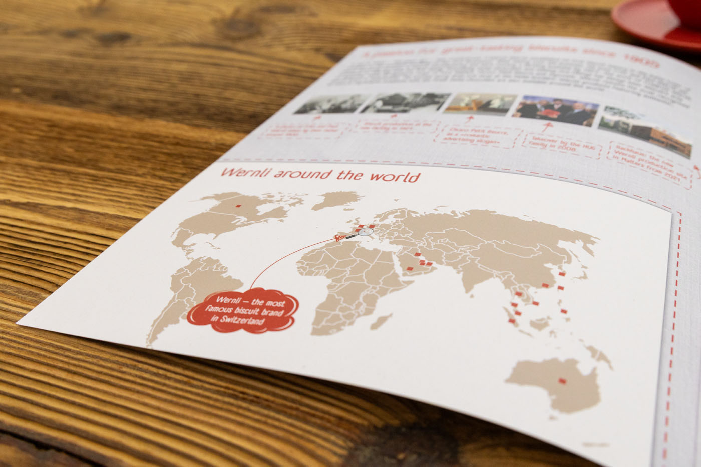 Nahaufnahme eines Blattes mit einer Weltkarte, auf der rot markierte Vertriebspunkte zu sehen sind. Die Überschrift lautet: „Wir sind überall auf der Welt“ und weist darauf hin, dass Verona die bekannteste Keksmarke der Schweiz ist. Das Blatt liegt auf einer Holzoberfläche.