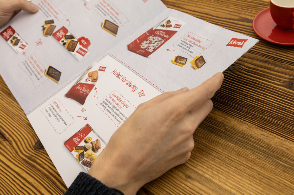 Eine Person hält eine weiße Broschüre mit Bildern und Beschreibungen verschiedener Snacks auf einem Holztisch. In der oberen rechten Ecke ist teilweise eine rote Kaffeetasse auf einer Untertasse zu sehen.