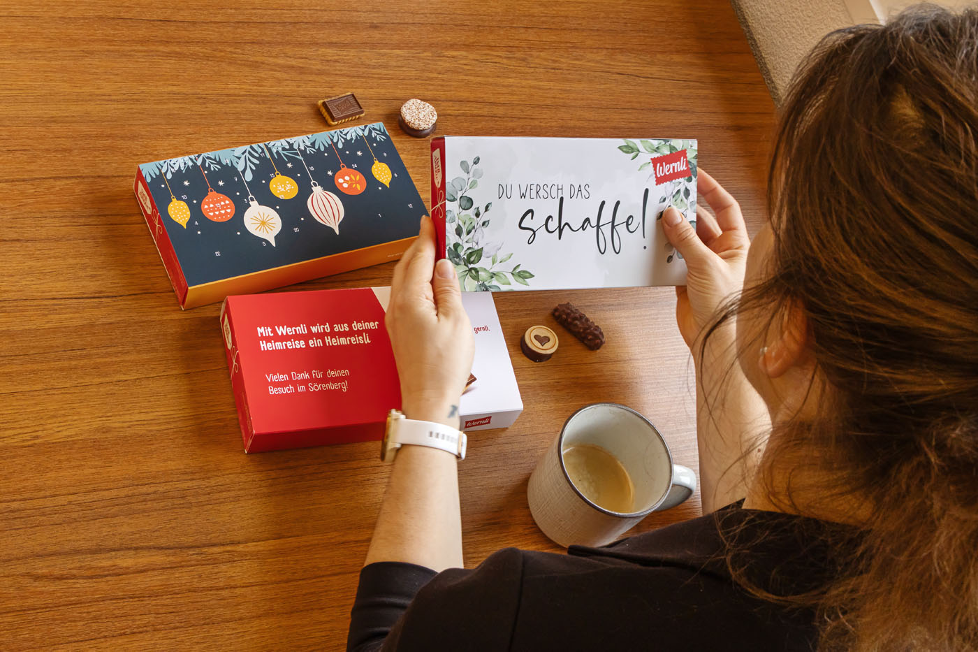 Eine Person hält eine Schachtel mit dekorativen Blättern und Text und sitzt an einem Tisch mit zwei weiteren bunten Schachteln, einer Tasse Kaffee und verschiedenen Snacks.