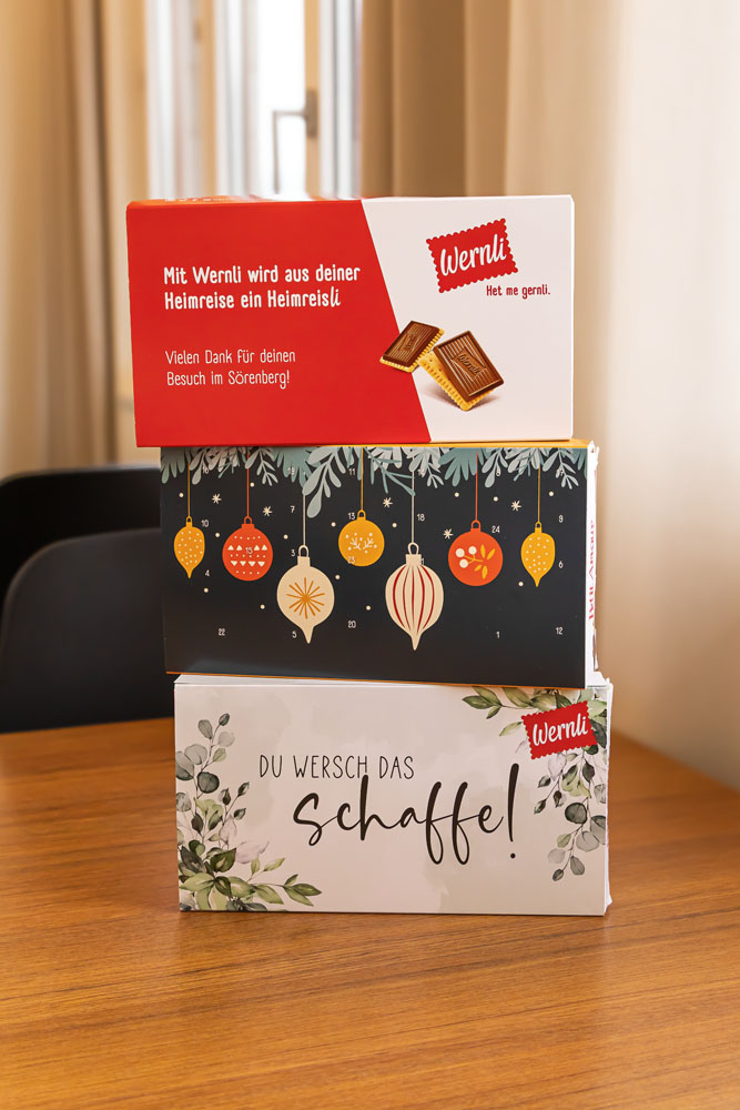 Ein Stapel aus drei bunten Schachteln auf einem Holztisch. Die obere Schachtel ist rot-weiß mit Schokoladenmotiven. Die mittlere Schachtel hat ein festliches Weihnachtsmotiv mit Ornamenten. Die untere Schachtel ist mit Blättern und dem Text „Du wersch das schaffe!“ verziert.