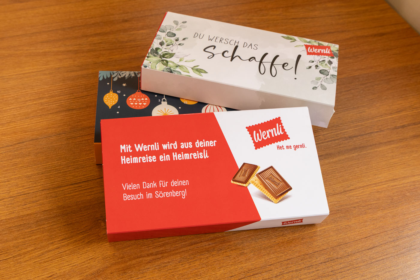 Zwei Schachteln Wernli-Kekse auf einem Holztisch. Eine Schachtel ist mit Weihnachtsschmuck verziert, die andere zeigt ein Bild von Schokoladenkeksen mit deutschem Text.