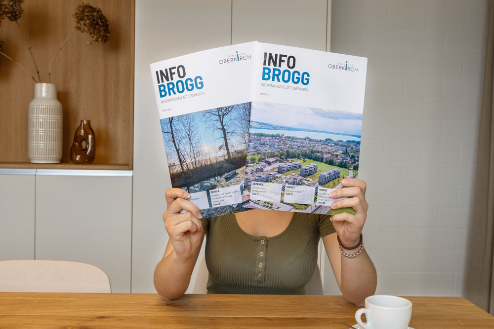 Eine Person sitzt an einem Tisch und hält zwei identische Broschüren mit dem Titel „INFO BROGG“ hoch. Diese zeigen Landschafts- und Gebäudebilder. Im Hintergrund ist ein Holzschrank zu sehen. Auf dem Tisch steht eine weiße Kaffeetasse.