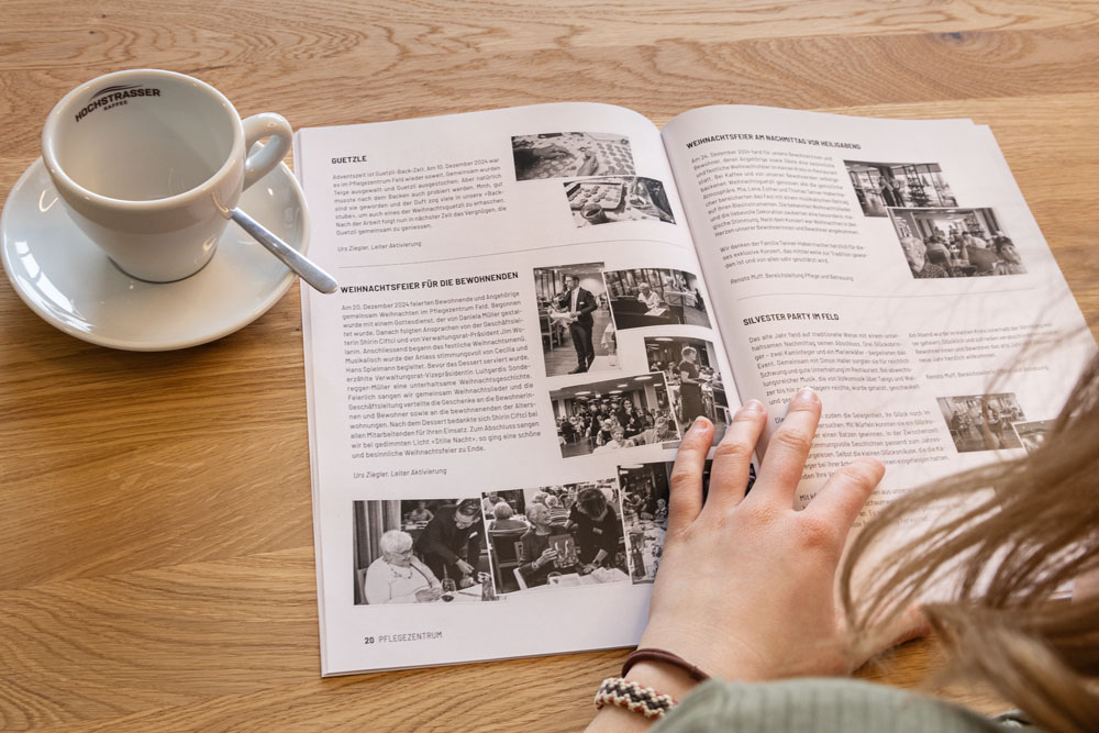 Eine Person liest eine Zeitschrift auf einem Holztisch. Ihre linke Hand liegt auf den aufgeschlagenen Seiten. Daneben steht eine weiße Kaffeetasse mit Untertasse und Löffel. Die Zeitschrift enthält Schwarz-Weiß-Fotos und Text.