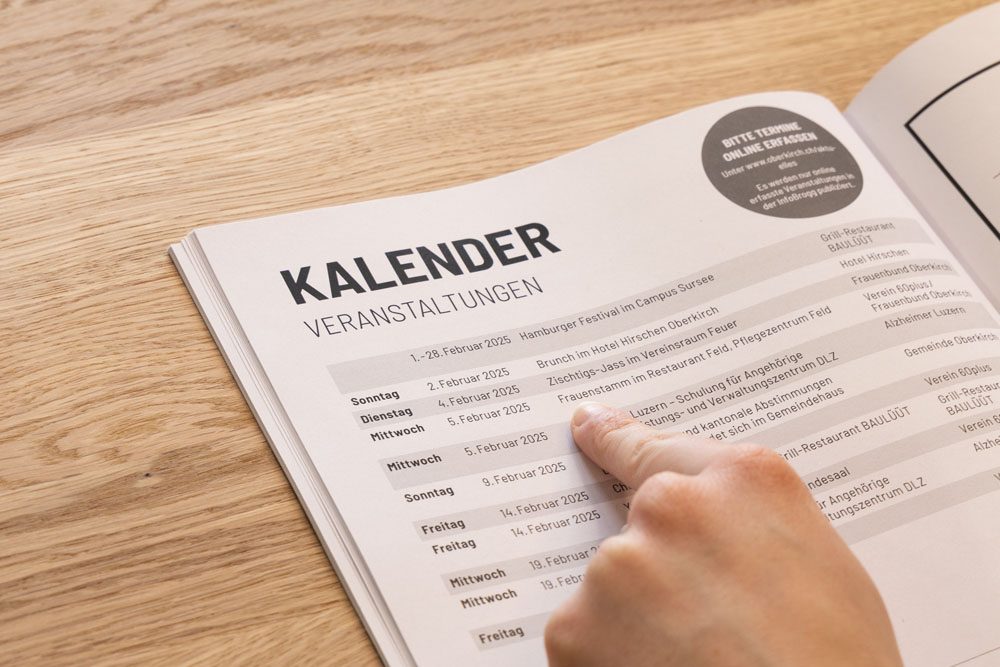 Eine Person zeigt auf eine Kalenderseite mit dem Titel „KALENDER VERANSTALTUNGEN“, auf der Veranstaltungsdaten und Details auf Deutsch aufgeführt sind. Das aufgeschlagene Heft liegt auf einer Holzoberfläche.