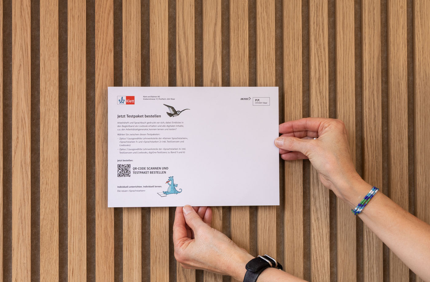 Zwei Hände halten einen weißen Umschlag vor einem Hintergrund aus Holzlatten. Der Umschlag ist mit Text, Logos und einem QR-Code bedruckt. Die Handgelenke der Person sind mit Armbändern und einer Uhr geschmückt.