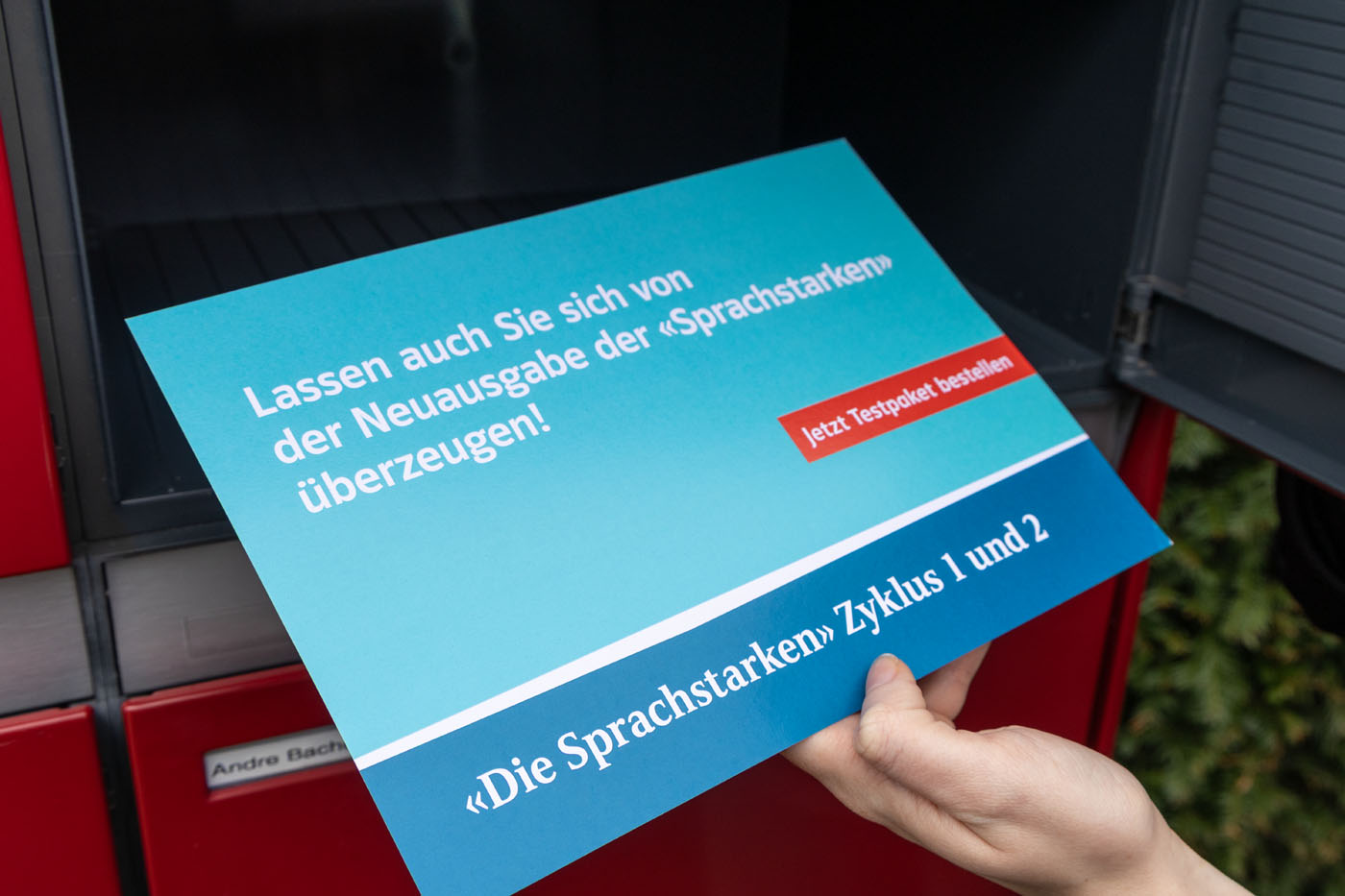 Eine Person hält ein blaues Schild vor einen roten Hintergrund. Auf dem Schild steht: „Lassen Sie sich auch von der Neuausgabe der ‚Sprachstarken‘ überzeugen!“ und „Die Sprachstarken Zyklus 1 und 2“.