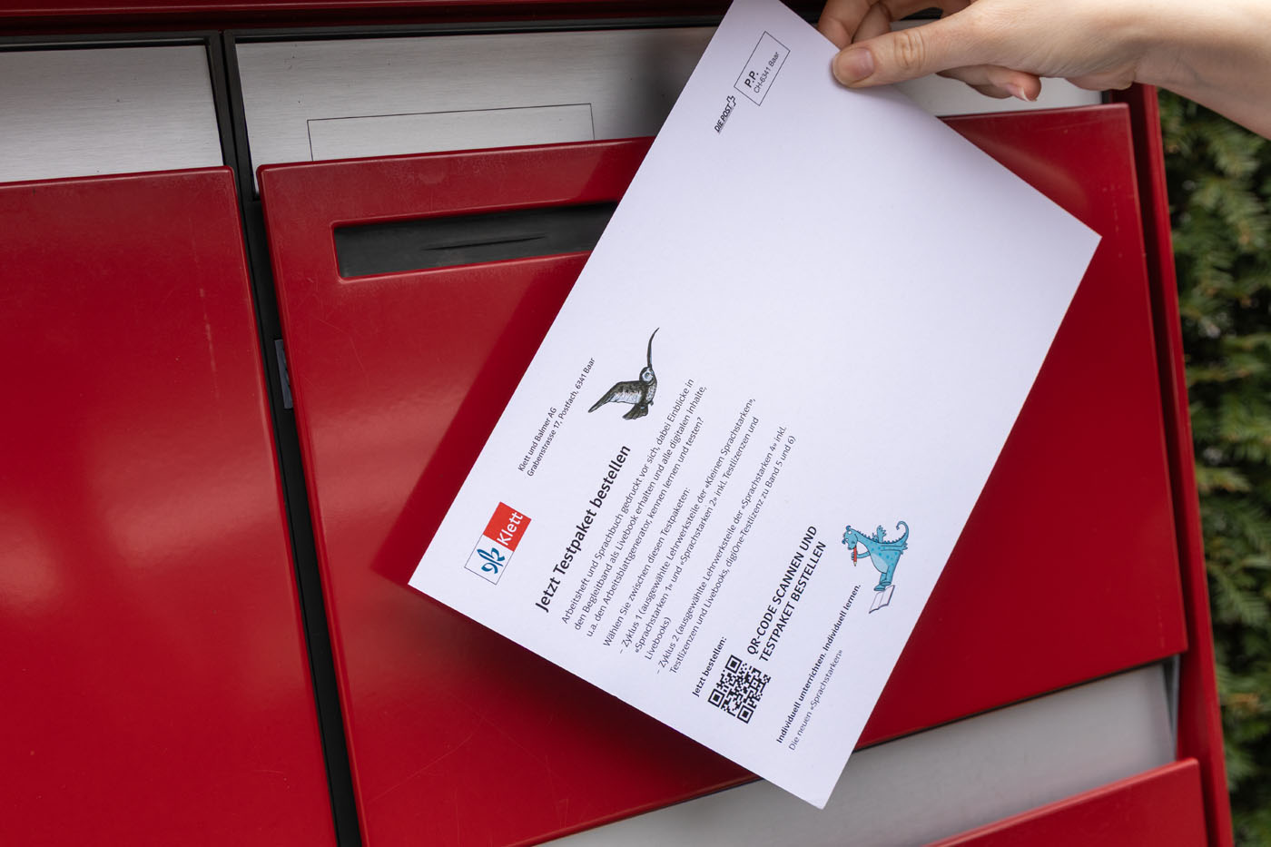 Eine Hand wirft einen Brief in einen roten Briefkasten. Der Brief enthält Text und Illustrationen, darunter eine Katze und eine Person in blauem Outfit. Ein QR-Code und Logos sind auf dem Brief zu sehen.