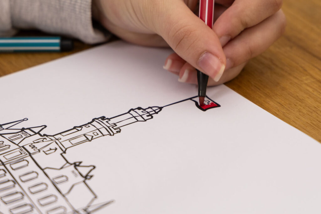 Nahaufnahme einer Hand, die mit einem roten Stift die Flagge auf einer Strichzeichnung eines Gebäudes mit Turm ausmalt. Das Papier liegt auf einer Holzoberfläche, daneben befindet sich ein blauer Stift.