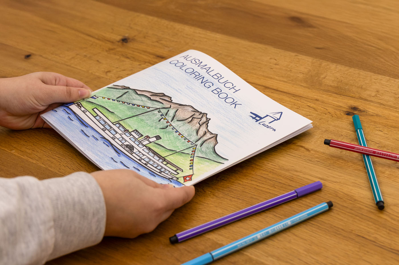 Hände halten ein zweisprachiges Malbuch mit dem Titel „Ausmalbuch Coloring Book“ mit der Zeichnung eines Bootes auf dem Wasser mit Bergen im Hintergrund. Farbige Stifte liegen verstreut auf einem Holztisch.