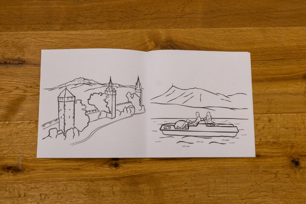 Ein aufgeschlagenes Malbuch auf einer Holzoberfläche zeigt zwei Seiten. Die linke Seite zeigt eine Burg mit Bergen. Die rechte Seite zeigt zwei Personen in einem Ruderboot auf einem See mit Bergen im Hintergrund.