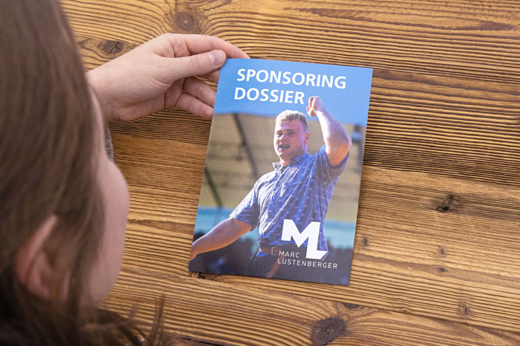 Eine Person hält eine Broschüre mit dem Titel „SPONSORING DOSSIER“ in der Hand. Darauf ist ein Mann in einem blauen Hemd abgebildet. Auf dem Cover sind außerdem die Initialen „ML“ und der Name „Marc Lustenberger“ zu sehen. Der Hintergrund ist eine Holzoberfläche.