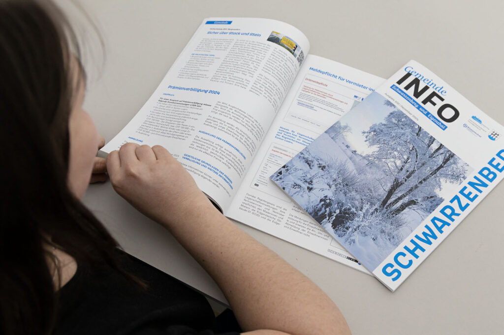 Person liest eine aufgeschlagene Zeitschrift auf einem Tisch. Auf dem Cover der anderen Zeitschrift „Gemeinde INFO“ ist eine verschneite Landschaft mit dem Wort „SCHWARZENBERG“ abgebildet. Die Szene suggeriert, den Inhalt zu lesen oder zu studieren.
