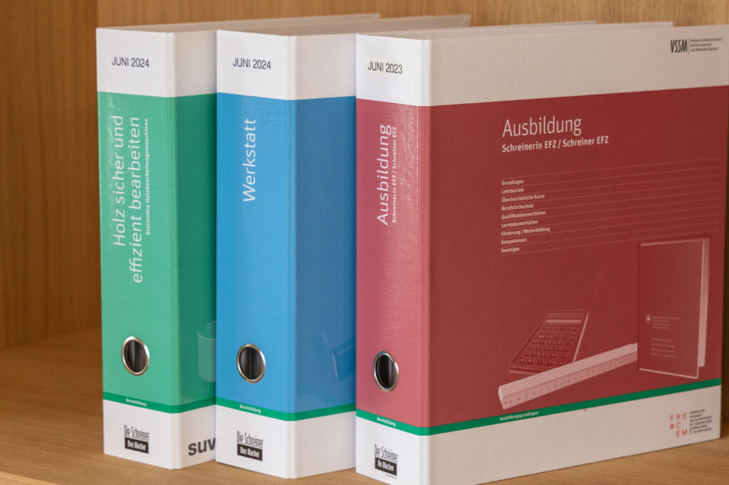 Drei Ordner auf einem Holzregal. Die Ordner tragen die grüne Aufschrift „Juni 2024“, die blaue Aufschrift „Werkstatt“ und die rote Aufschrift „Ausbildung“ mit der Jahreszahl „Juni 2023“. Jeder Ordner enthält gedruckte Texte und Grafiken zum Thema Holzbearbeitung.