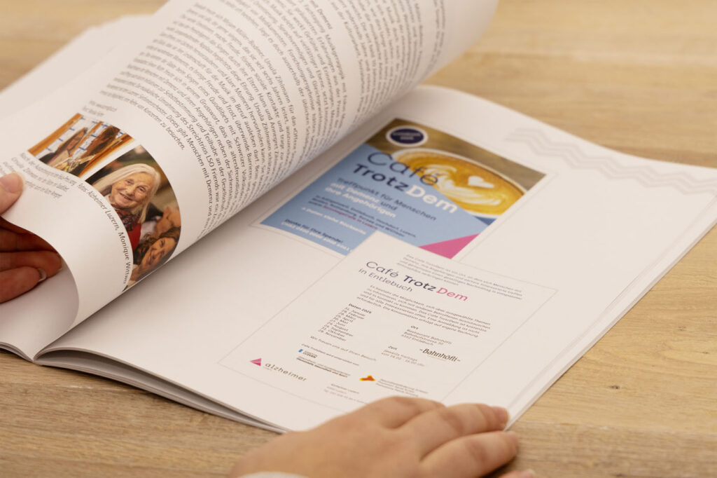 Eine Person blättert in einer Zeitschrift. Der Fokus liegt auf einer Seite mit einem Flyer mit dem Titel „Café Trotz Dem“. Der Flyer enthält Text und Grafiken auf hellem Hintergrund. Die Zeitschrift liegt aufgeschlagen auf einem Holztisch, Hände blättern sichtbar um.
