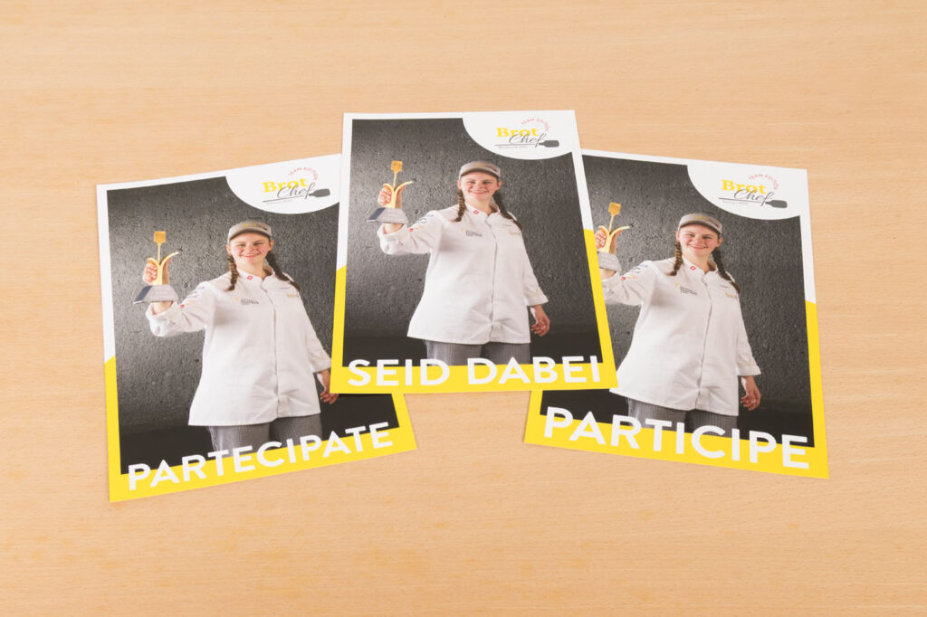 Drei Flyer zeigen eine lächelnde Frau in Kochjacke und -mütze mit einem Pfannenwender in der Hand. Jeder Flyer trägt unten die Worte „PARTICIPATE“, „SEID DABEI“ und „PARTICIPE“. Der Hintergrund ist aus hellem Holz.