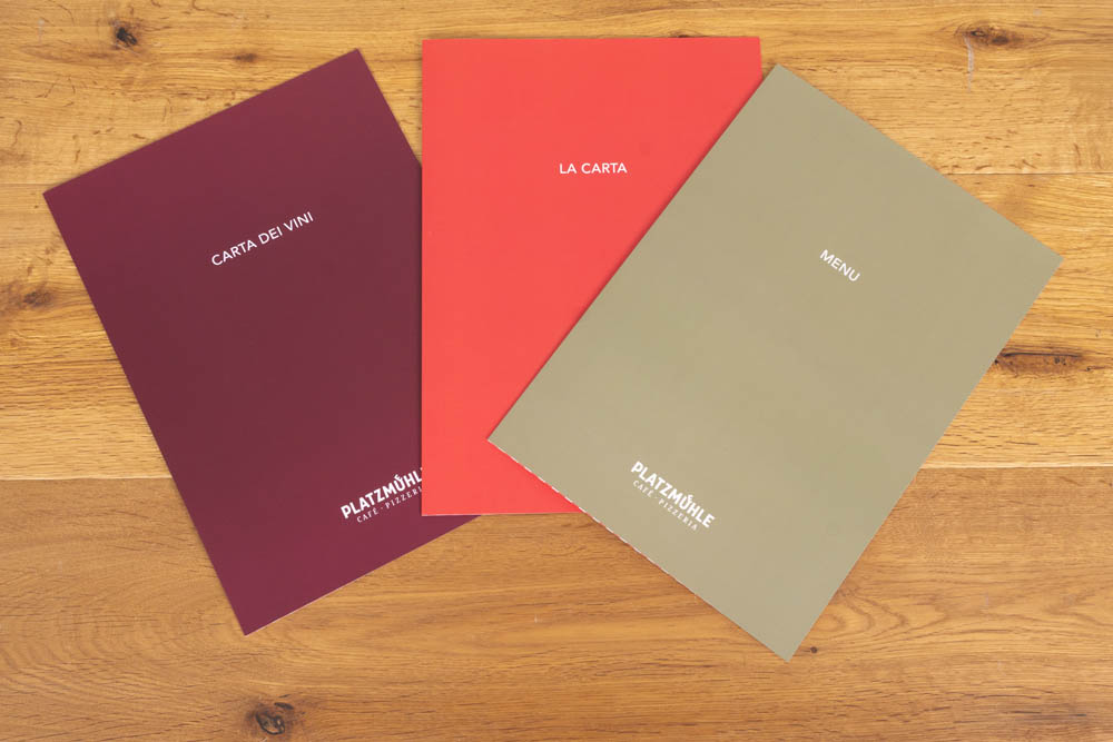 Auf einer Holzplatte liegen drei Speisekarten: eine weinrote mit der Aufschrift "CARTA DEI VINI", eine rote mit der Aufschrift "LA CARTA" und eine olivgrüne mit der Aufschrift "MENU", jeweils mit der Aufschrift "PLATZMÜHLE" am unteren Rand.