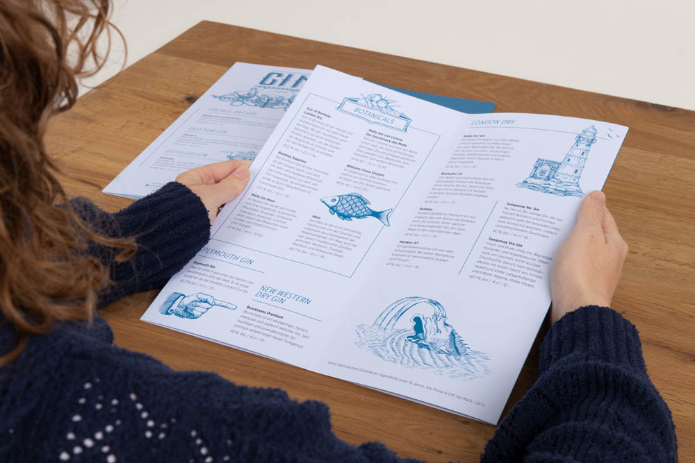 Eine Person in einem marineblauen Pullover sitzt an einem Holztisch und liest eine weiß-blau illustrierte Speisekarte mit Gin-Optionen, auf der ein Fisch, eine Welle und ein Leuchtturm abgebildet sind.