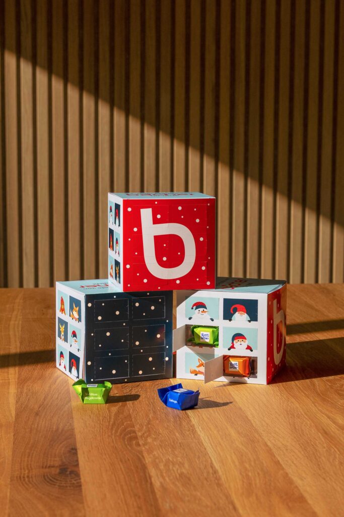Auf einem Holztisch stehen drei farbenfrohe, festlich gestaltete Adventskalenderboxen mit kleinen Schubladen, die einzeln verpackte Leckereien enthalten. Das Sonnenlicht wirft Schatten auf die holzgetäfelte Wand hinter ihnen.