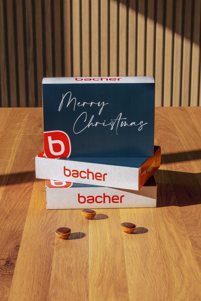 Drei gestapelte Geschenkschachteln, zwei mit der Aufschrift "bacher" in roter Farbe und eine blaue Schachtel obenauf mit "Frohe Weihnachten" in weißer Schrift, stehen auf einem Holztisch mit drei kleinen Leckereien davor.