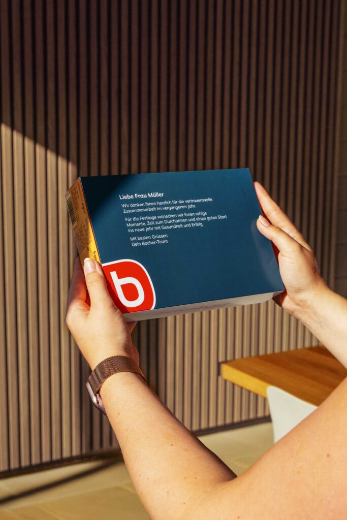 Eine Person, die eine blaue Schachtel mit deutschem Text und einem roten "b"-Logo in der Hand hält, steht in einem gut beleuchteten Raum mit Holzverkleidung und einem Tisch im Hintergrund.