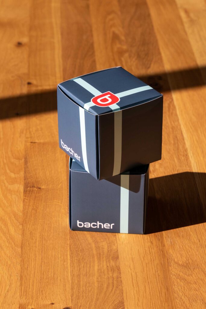 Zwei dunkelblaue würfelförmige Schachteln mit der Aufschrift "bacher" sind auf einer Holzfläche gestapelt, wobei das Sonnenlicht Schatten auf den Boden wirft. Die oberste Schachtel ist mit einem stilisierten rot-weißen Logo versehen.