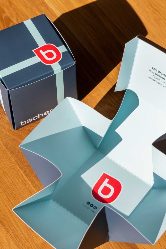 Zwei Schachteln auf einer Holzoberfläche: eine ist geschlossen und dunkelblau mit einem hellblauen Streifen und einem roten "b"-Logo; die andere ist offen und zeigt ein hellblaues Inneres, das ebenfalls das rote "b"-Logo und einen gedruckten Text aufweist.