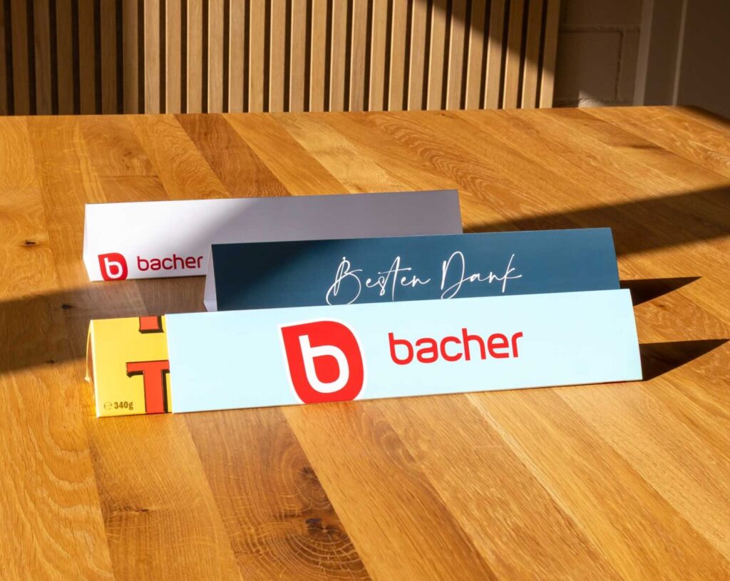 Zwei blaue Papiertischzelte mit dem roten "bacher"-Logo und weißem Text stehen auf einem Holztisch im Sonnenlicht. Im Hintergrund sind vertikale Holzwandpaneele und eine geschlossene Tür zu sehen.